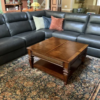 Contemporary dark gray leather sectional sofa, accompanied by a wooden coffee table with woven rattan aprons and a floral pattern area rug.