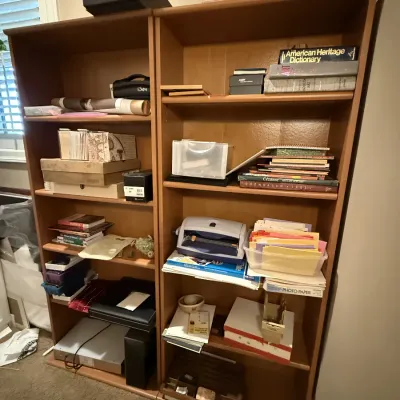 Pair of tall light oak bookcases filled with books, crafting supplies, office materials, and miscellaneous decor items.