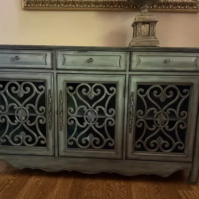 Distressed blue-green painted sideboard featuring three upper drawers and three cabinet doors with ornate wrought-metal scrollwork overlays.