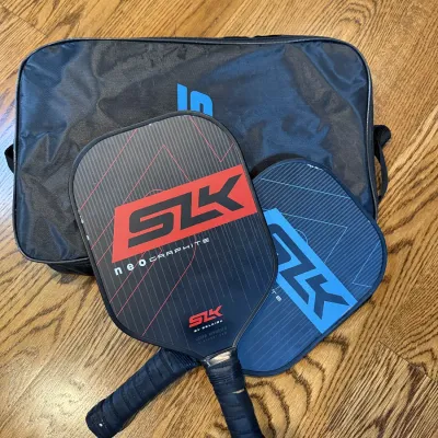 Pair of SLK by Selkirk Neo Graphite pickleball paddles in red and blue, shown with a black carrying bag.