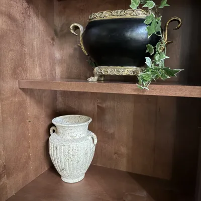 Black urn with gilded handles and base holding faux ivy, alongside a textured cream-colored urn.