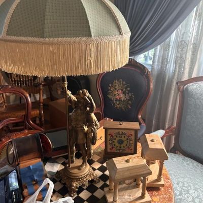 Sale Photo Thumbnail #35: Vintage brass table lamp featuring a sculpted figure holding a staff, paired with a fringed green fabric dome shade, shown alongside various decorative clocks.