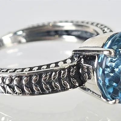 Sterling silver ring featuring a round-cut blue topaz gemstone set in a four-prong mount, accented by a textured, oxidized wheat-pattern...