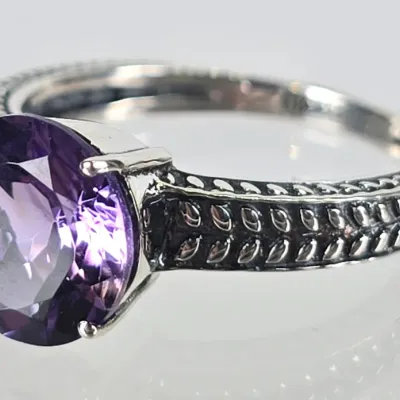 Sterling silver ring featuring a round-cut purple amethyst gemstone in a four-prong setting, accented by a textured, leaf-patterned band.