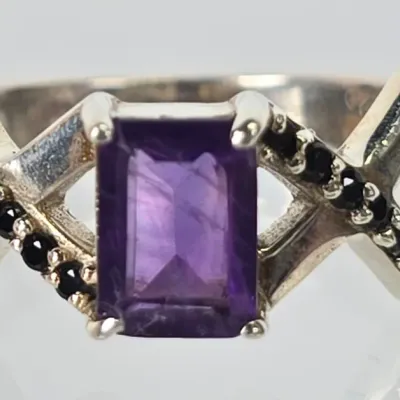 Sterling silver ring featuring an emerald-cut purple amethyst center stone flanked by crisscrossing bands accented with small black...