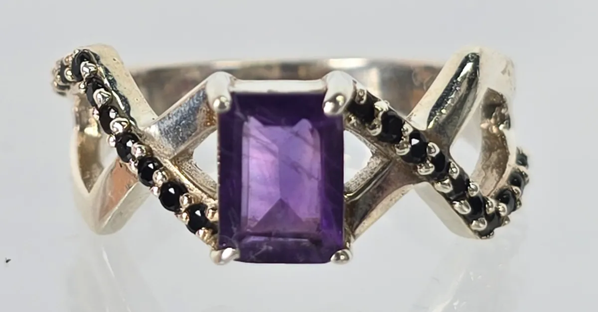Sterling silver ring featuring an emerald-cut purple amethyst center stone flanked by crisscrossing bands accented with small black...