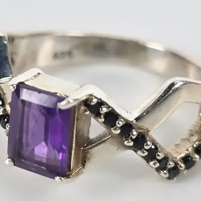 Sterling silver ring featuring an emerald-cut purple amethyst center stone flanked by black spinel accents on a crisscross band.