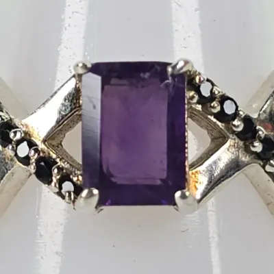 Sterling silver ring featuring an emerald-cut purple amethyst center stone, flanked by criss-cross bands accented with small black...
