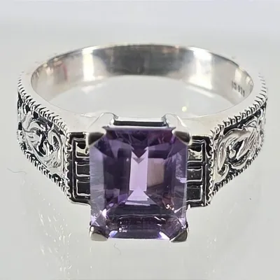 Sterling silver ring featuring an emerald-cut amethyst gemstone, accented by ornate filigree and textured detailing along the band.