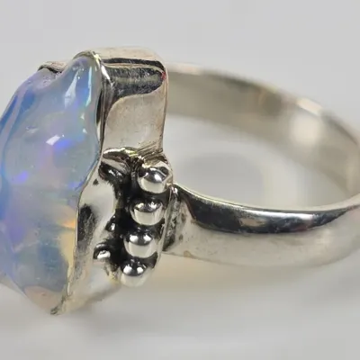 Sterling silver ring featuring a large, raw-cut opal stone with iridescent blue and purple play-of-color, accented by beaded metalwork on...