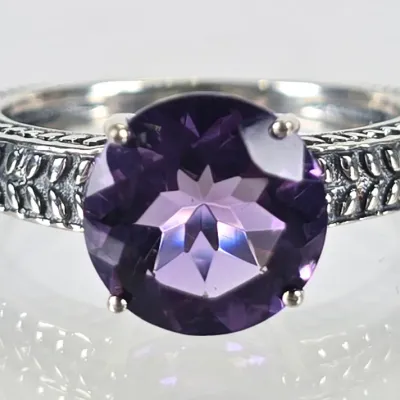 Sterling silver ring featuring a round-cut purple amethyst gemstone secured in a four-prong setting, accented by a textured,...