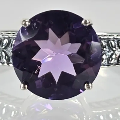 Sterling silver ring featuring a round-cut purple amethyst gemstone with a starburst facet pattern, set on a band with textured leaf-like...