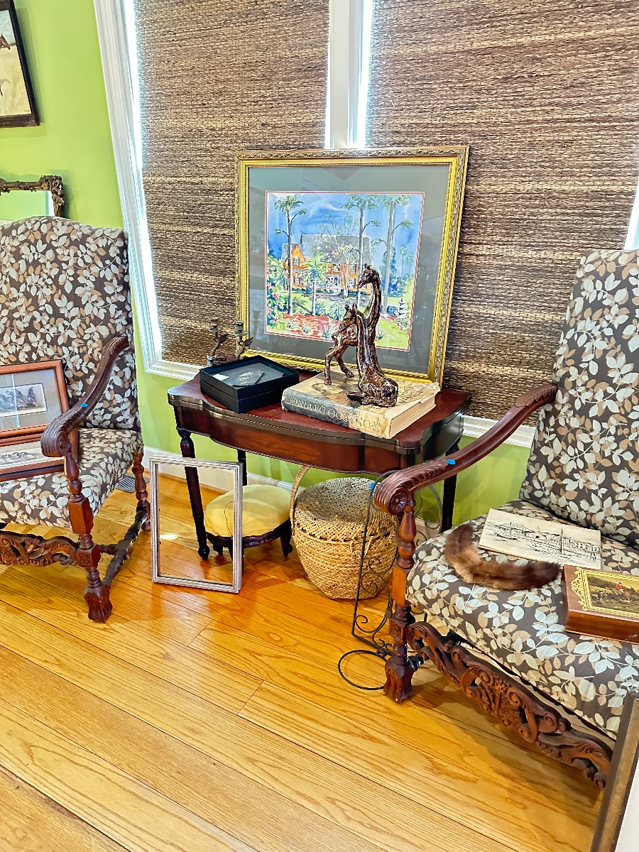 Pair of ornate carved wood armchairs with leaf-patterned upholstery, flanking a mahogany console table displaying framed artwork, a...