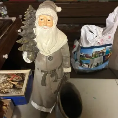 Vintage-style Santa Claus figurine in a grey coat, holding a small evergreen tree and a sack that functions as a small planter or holder.