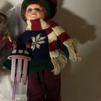 Vintage porcelain boy doll dressed in winter attire, featuring a knit sweater, striped scarf, and matching hat while holding a miniature...