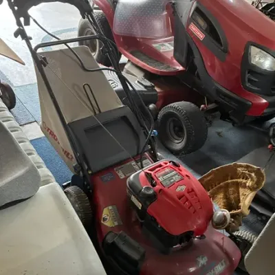 Troy-Bilt walk-behind lawn mower with rear bagger, shown alongside a red Craftsman riding lawn mower in a garage setting.