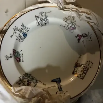 Vintage porcelain plate featuring whimsical illustrations of figures celebrating, accented with a gold-gilded rim and wrapped in...