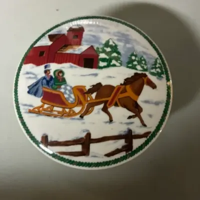 Vintage round ceramic trinket box lid featuring a winter scene of a horse-drawn sleigh with a couple, a red barn, and evergreen trees.