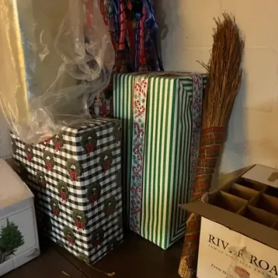 Assorted wrapped Christmas gifts and holiday decorations, including a decorative cinnamon broom, stored on a shelf.