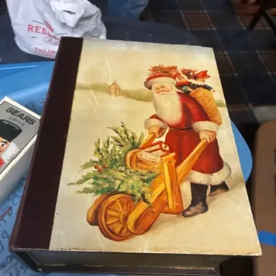 Vintage book-shaped Christmas storage box featuring an illustration of Santa Claus pushing a wheelbarrow filled with a Christmas tree and...