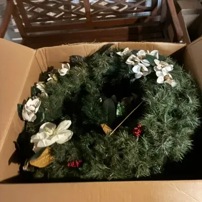 Large artificial evergreen Christmas wreath decorated with white magnolia blossoms and red berries, stored in a cardboard box.