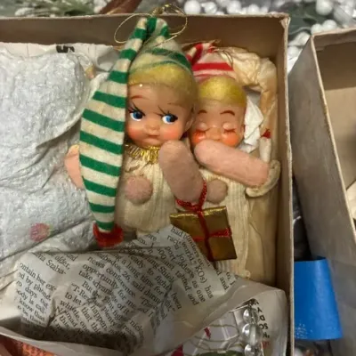 Pair of vintage mid-century Christmas elf ornaments with felt hats, plastic faces, and fabric bodies, nestled in a cardboard storage box.