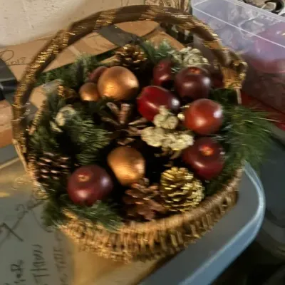 Festive holiday basket arrangement featuring faux apples, gilded pinecones, and evergreen sprigs in a woven basket with a wrapped handle.