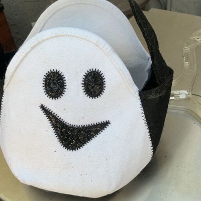Plush felt Halloween trick-or-treat basket featuring a smiling white ghost face with black glitter eyes and mouth.