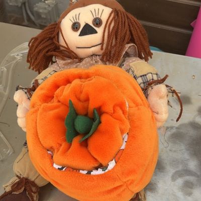 Plush scarecrow doll holding a large soft pumpkin, featuring yarn hair and a patchwork hat for autumn decor.