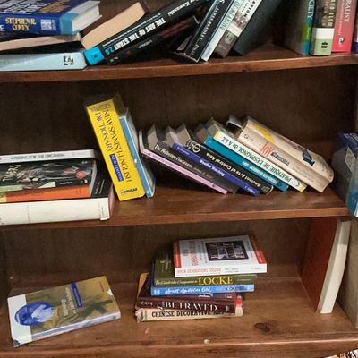 A collection of non-fiction and educational books, including dictionaries, history, and cookbooks, arranged on a wooden bookshelf.