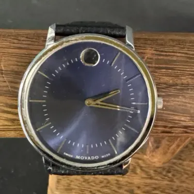 Vintage Movado Swiss-made wristwatch featuring a dark blue dial with gold-tone hands, a signature dot at 12 o'clock, and a black...