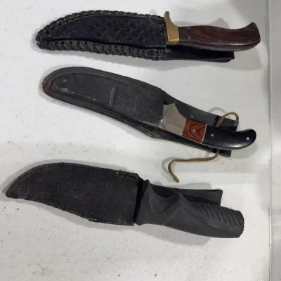 Three fixed-blade hunting knives, each housed in its respective black sheath, featuring various handle styles including wood grain and...