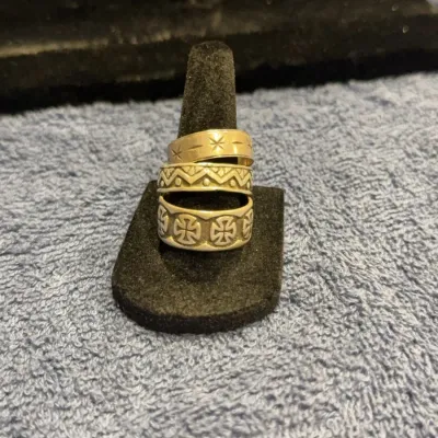 Three silver-tone band rings with engraved patterns, including starburst, geometric, and Maltese cross designs, displayed on a black...