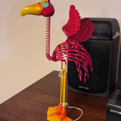 Whimsical metal flamingo bobble figure, featuring a coiled spring neck, vibrant pink and yellow painted finish, and stylized wings,...