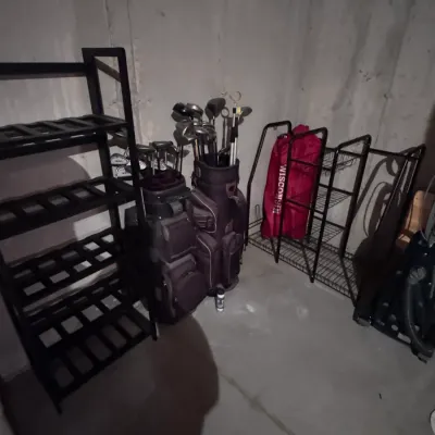 Two golf bags filled with clubs, including a set of Ping irons, alongside a black wooden shelving unit and a wire storage rack in a...