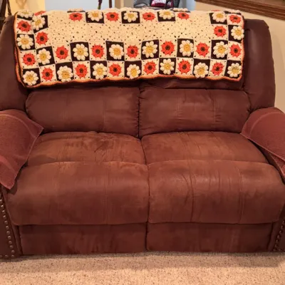 Brown microfiber reclining loveseat featuring nailhead trim, draped with a vintage handmade crochet granny square blanket in autumn tones.