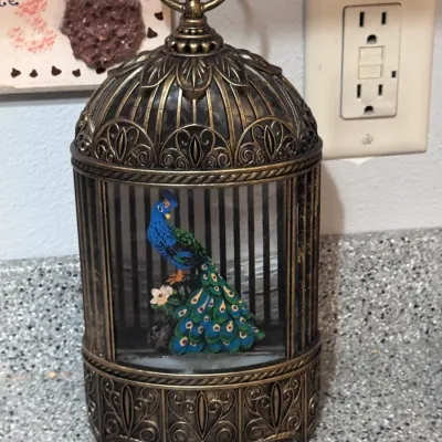 Vintage-style decorative birdcage lantern featuring an ornate bronze-finished plastic frame and a colorful peacock figurine inside,...