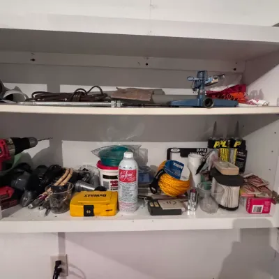 Assorted garage and workshop supplies on white shelving, featuring a DeWalt drill bit case, Sea Foam motor treatment, a yellow rope...