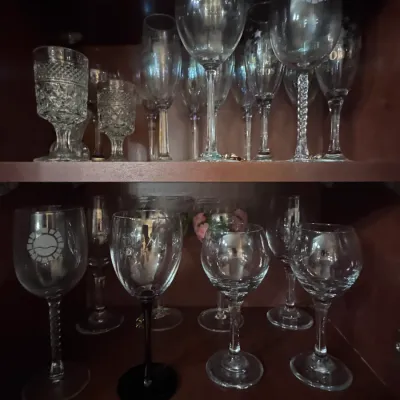 Assorted glassware collection, featuring crystal wine goblets, champagne flutes, and etched souvenir glasses displayed on wooden cabinet...