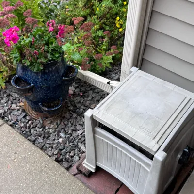 Suncast plastic hose reel box in light gray, alongside a blue glazed ceramic strawberry planter with pink geraniums.