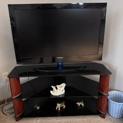 Samsung flat-screen television on a modern corner TV stand featuring black tempered glass shelves and cherry-finished wood support pillars.