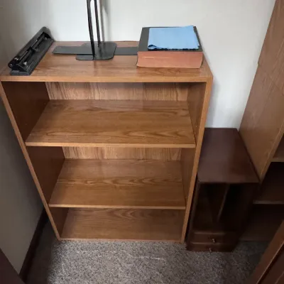 Three-shelf oak veneer bookcase featuring adjustable shelves, shown holding office supplies including a heavy-duty hole punch and bookends.