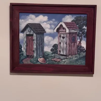 Original folk art painting depicting two rustic outhouses in a grassy field under a cloudy sky, housed in a simple reddish-brown wooden...