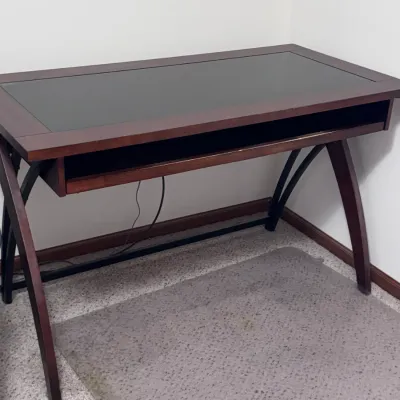 Modern wooden writing desk featuring a dark glass-inlaid top, a pull-out keyboard tray, and curved wooden legs with black metal support...
