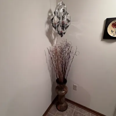 Mid-century modern style chrome ribbon hanging candle holder chandelier, paired with a tall ceramic floor vase holding decorative dried...
