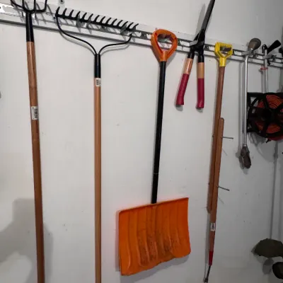 Assorted garden and lawn tools hanging on a wall organizer, including a bow rake, a thatch rake, an orange plastic snow shovel, hedge...