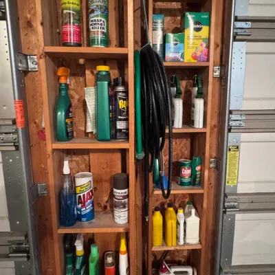 Assorted garage and garden chemicals stored on a wooden shelving unit, featuring brands like Miracle-Gro, Ortho, Spectracide, and...