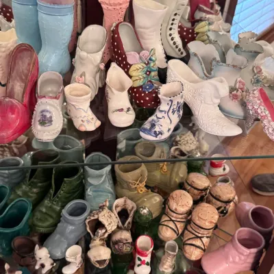 Collection of vintage ceramic shoe figurines displayed on glass shelves, featuring various styles including Victorian boots, high heels,...