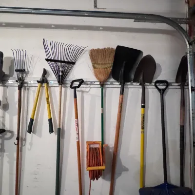 Assorted garden and yard tools hanging on a wall organizer, including a True Temper fiberglass shovel, Ames leaf rakes, loppers, a straw...