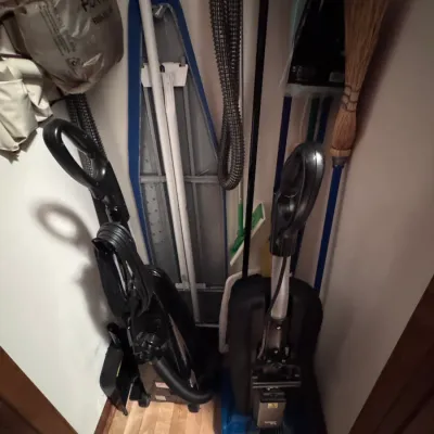 Assorted household cleaning supplies stored in a closet, including two upright vacuum cleaners, a folding ironing board with a blue...